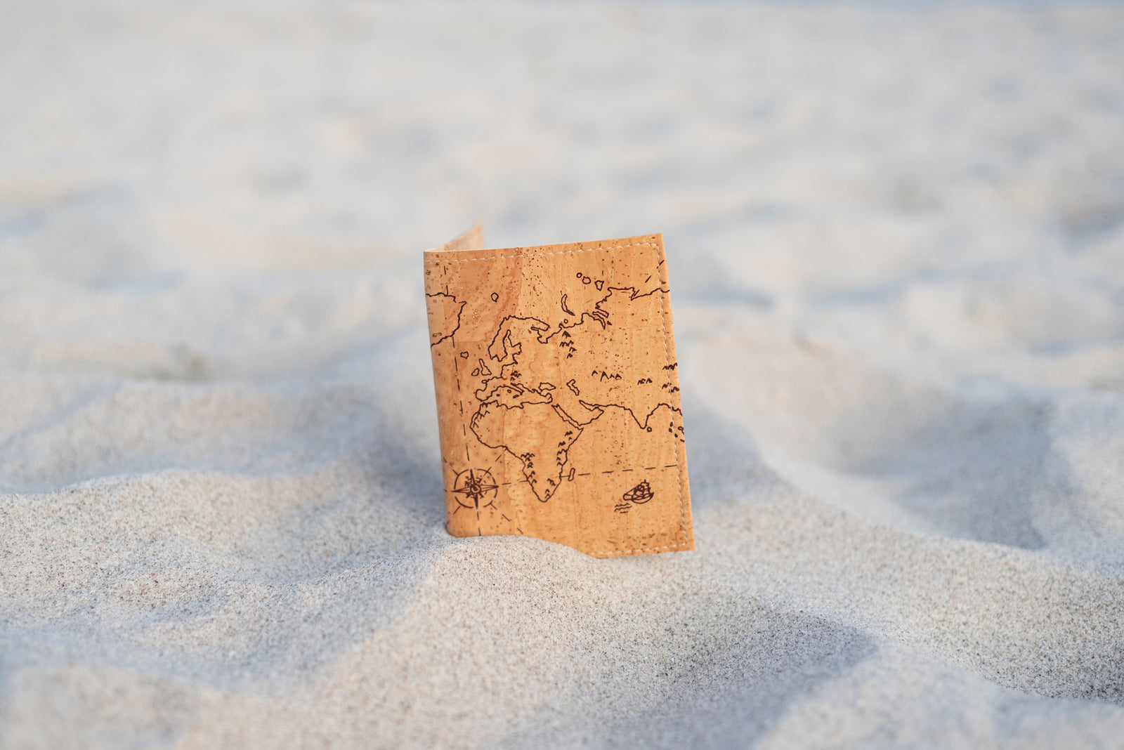 Reisepasshülle aus Kork mit Weltkarten-Motiv steht im Sand an einem Strand
