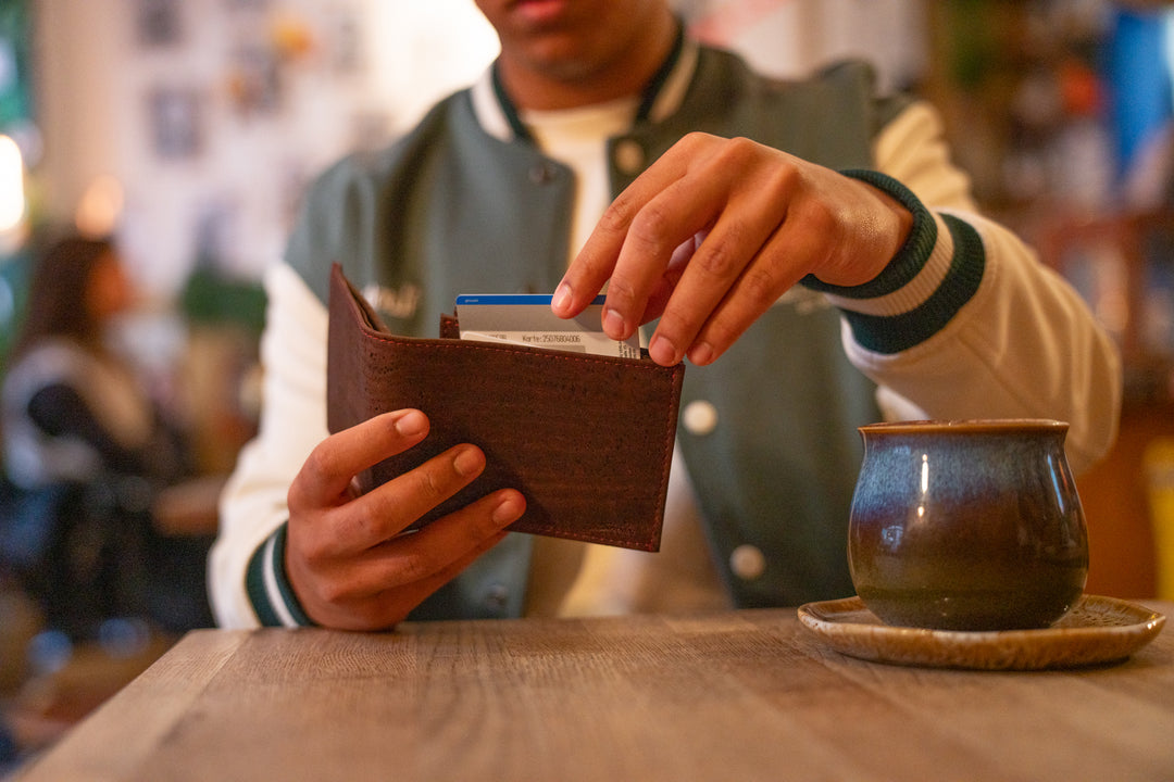 Junger Mann sitzt an Tisch in Cafe und zieht Geldkarte aus geöffnetem Kork Geldbeutel, Kaffeetasse auf Holztisch