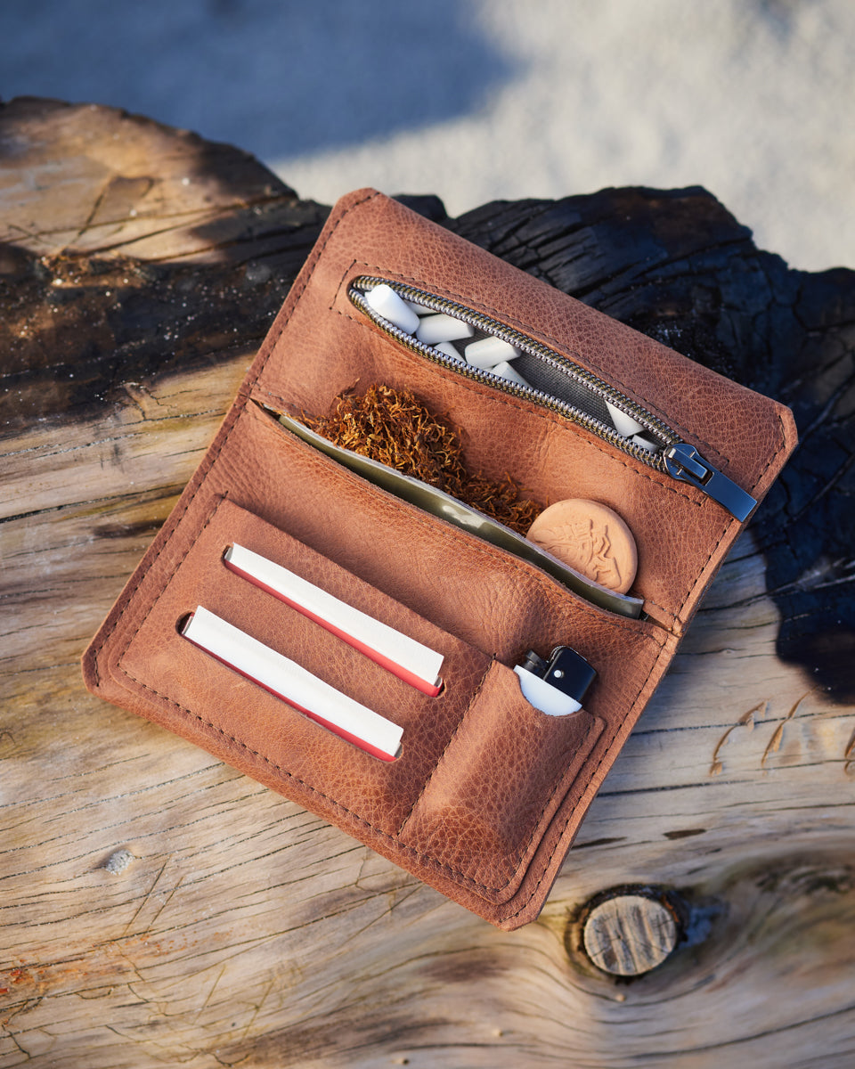Braune Ledertabaktasche mit Inhalt liegt geöffnet auf einem größen Holzstück am Strand