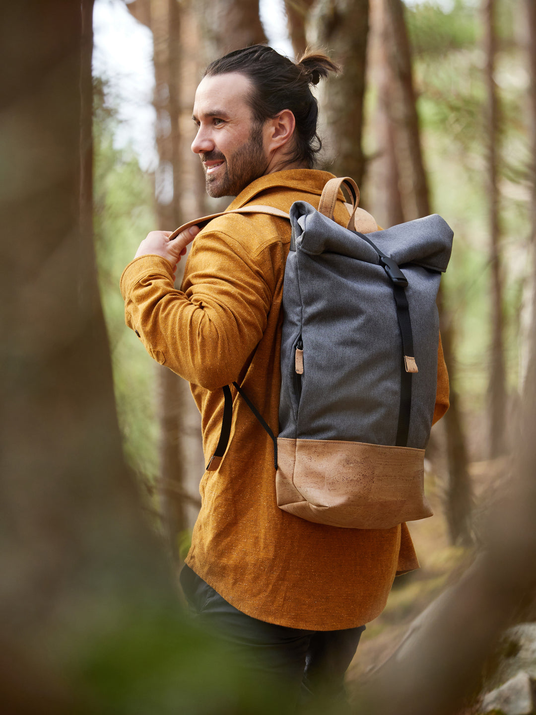 Junger Mann im Wald trägt lächelnd grauen Rucksack mit hellem Korkboden über der linken Schulter