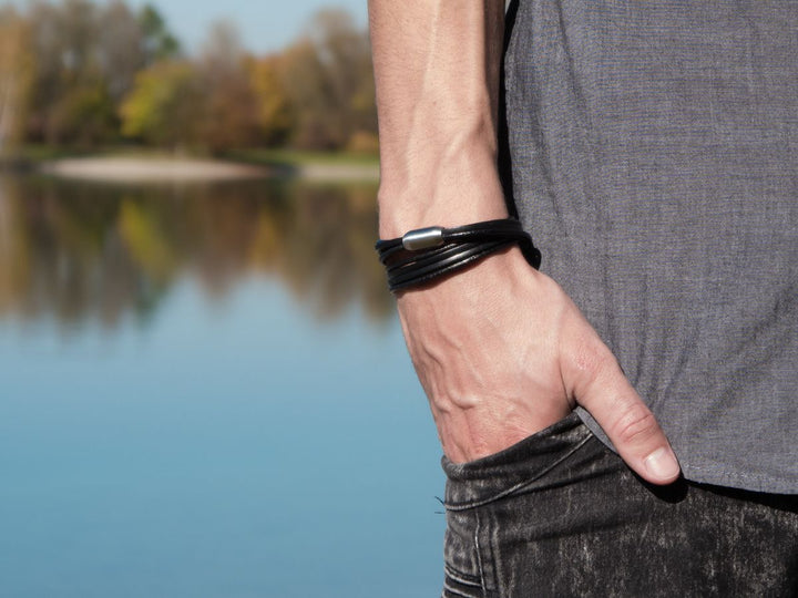 Mann mit schwarzem Wickelarmband aus Leder am rechten Handgelenk posiert vor einem See