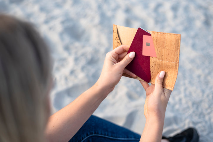 Frau sitzt am Strand und zieht Reisepass aus Reisepasshülle aus hellem Kork #color_weltkarte