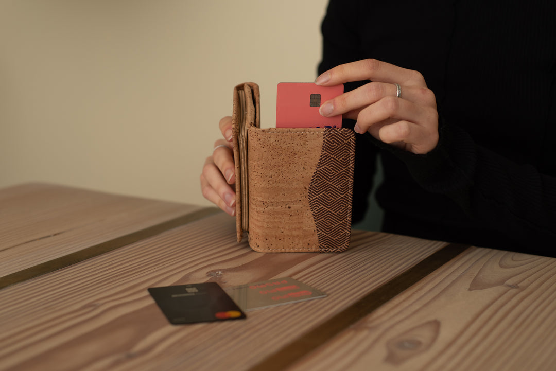 Frau in schwarzem Pullover sitzt an Holztisch und zieht Geldkarte aus hellem Kork Geldbeutel mit Wellenmuster #color_raizes