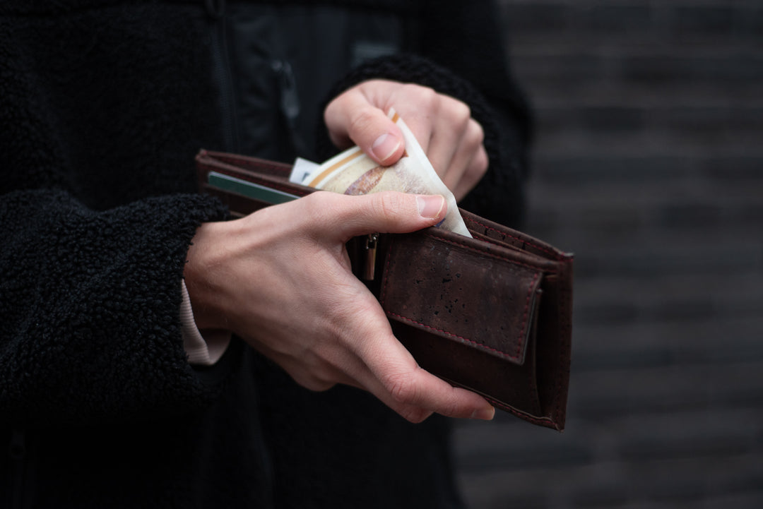 Person in schwarzem Wintermantel zieht Geldscheine aus dunkelbraunem Kork Geldbeutel