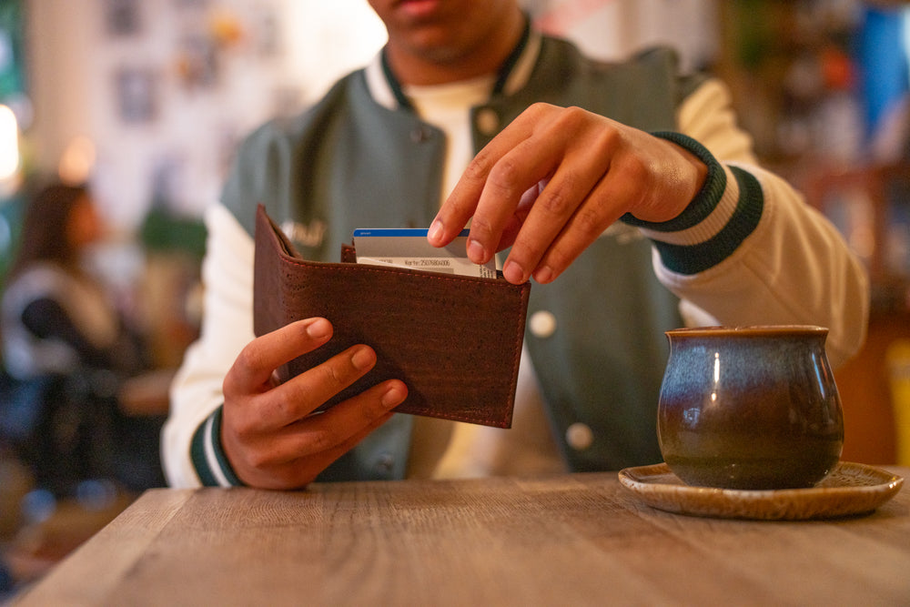Junger Mann sitzt an Tisch in Cafe und zieht Geldkarte aus geöffnetem Kork Geldbeutel, Kaffeetasse auf Holztisch
