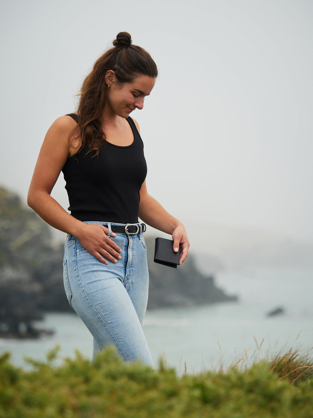 Junge Frau geht mit schwarzem Ledergeldbeutel in der linken Hand fröhlich am Meer spazieren