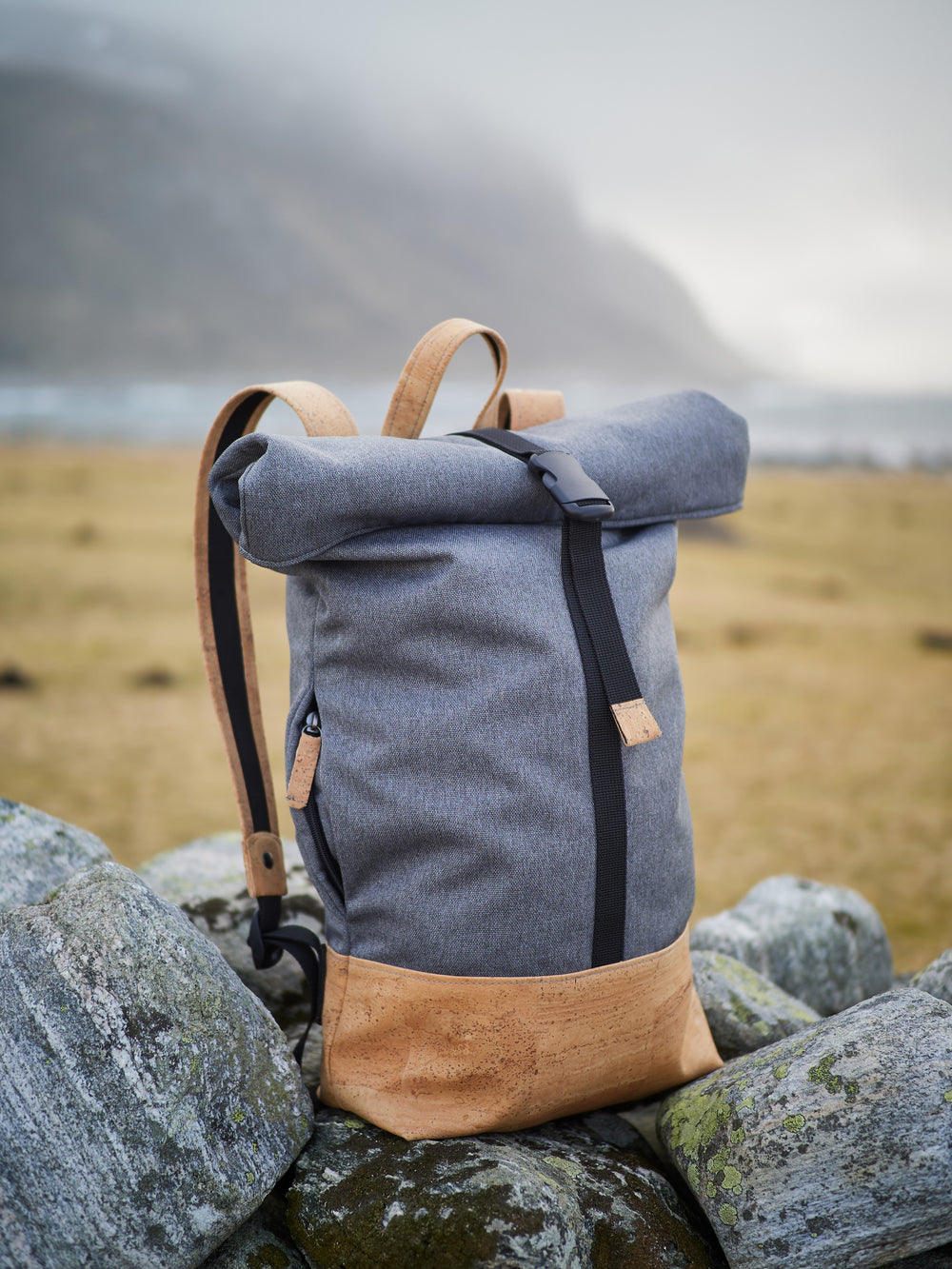 Grauer Rolltop Rucksack mit hellem Kork Boden steht auf Steinen mit Aussicht auf das Meer im Hintergrund